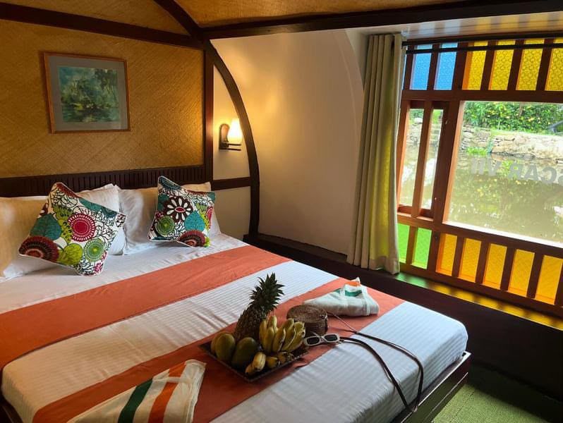 Double bed with white linen and patterned cushions beside a coloured-glass window looking onto the water